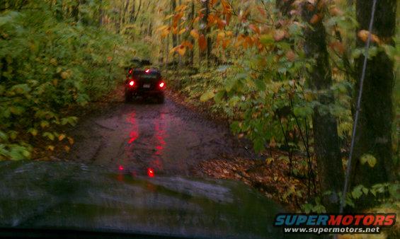 Dark SUV with red taillights on a muddy, wet road in autumn woods