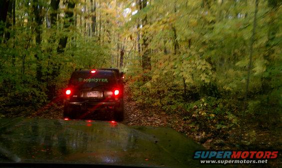 Black SUV with Monster Energy sticker on rear window, driving on a dirt road in a forest