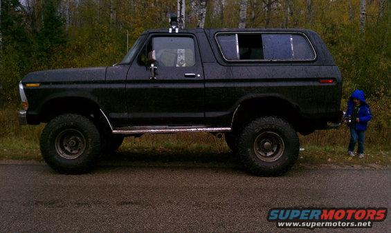 Black 1978 Ford Bronco parked on the side of the road, lifted with off-road tires. A child stands near the rear