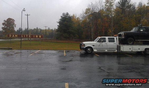 White flatbed truck carrying a black pickup truck, parked on a wet lot near a sign for Dam Site Inn