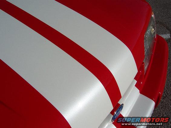 Red car hood with white racing stripes, showing part of the headlight and a Ford emblem