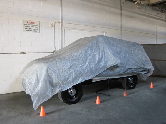Vehicle covered with a gray tarp, parked indoors with orange cones on the floor