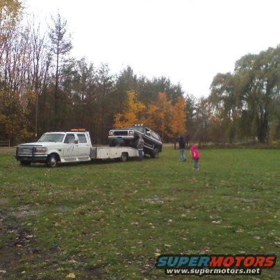 White tow truck with a dark SUV on its trailer, two people stand nearby in a grassy field
