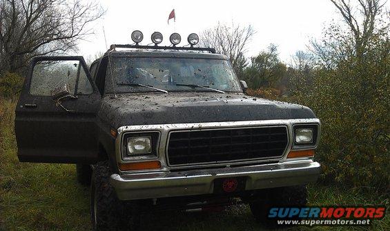 Mud-covered 1979 Ford Bronco with driver's door open, parked in a grassy, wooded area