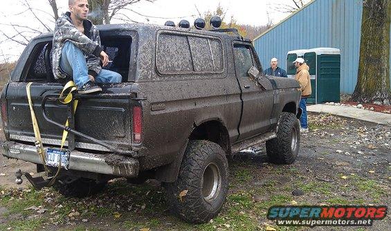 Mud-caked SUV, rear view, with a person sitting in the open tailgate and a spare tire mount