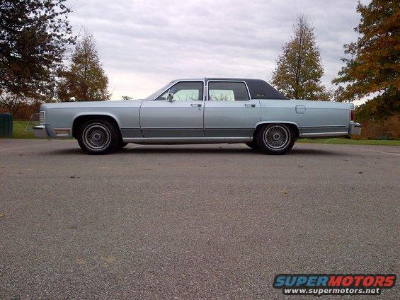 Light blue Lincoln Town Car parked on asphalt, trees in background