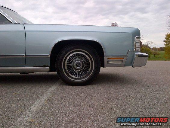 Light blue sedan's front quarter panel, fender, and wire wheel. Chrome bumper and amber marker light visible