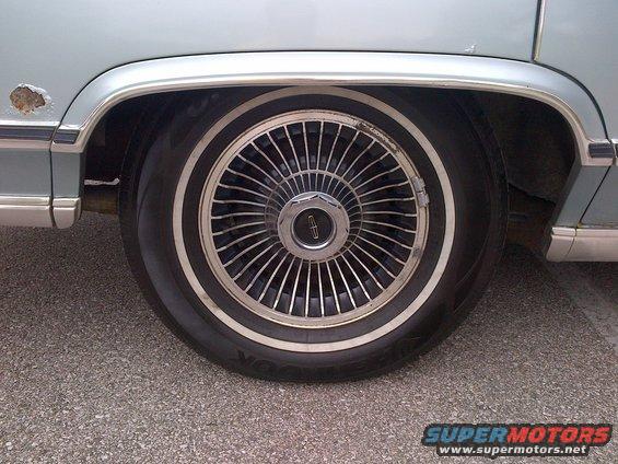 Light blue car wheel with chrome hubcap and whitewall tire, showing rust on fender