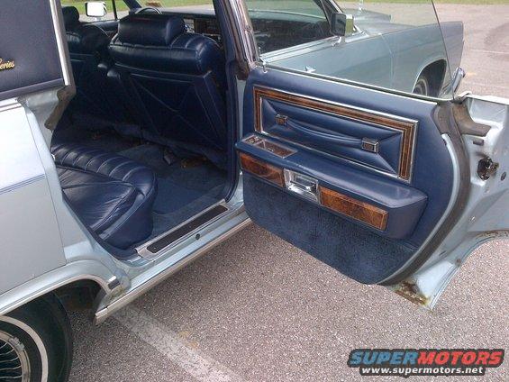 Light blue Lincoln Continental interior with blue leather seats and wood trim on the door panel. Rust is visible on the lower door edge