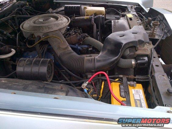 Engine bay showing air cleaner assembly, hoses, and yellow Optima battery with red positive terminal cable