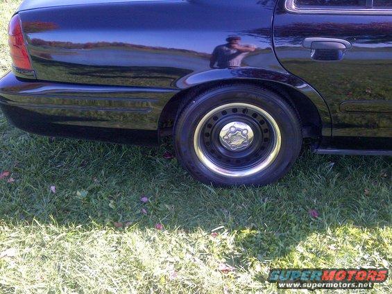 Rear quarter panel and wheel of a black sedan parked on grass. The wheel has a chrome hubcap