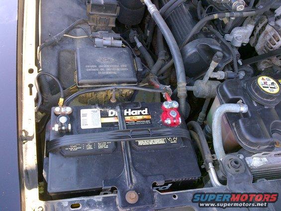 Car battery with red terminal connectors in engine bay. Ford logo visible on a black box