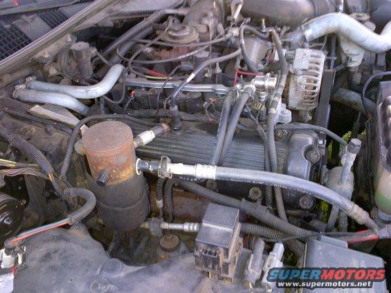 Engine bay with various hoses, wires, and engine components visible. A black fuse box is in the lower right
