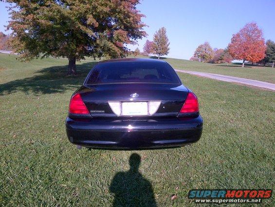 Black sedan parked on grass with trees in background. License plate area is blank