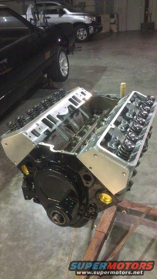 V8 engine block with aluminum valve covers and exposed pushrods, resting on a stand. A black car and a silver truck are visible in the background