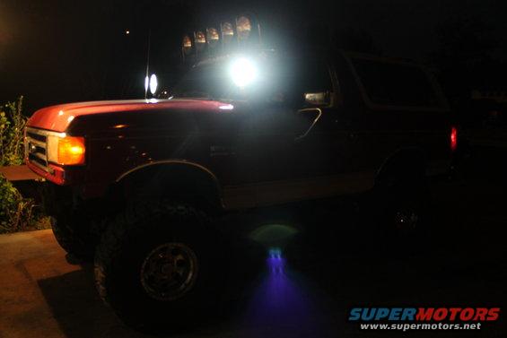 Red 1990 Ford Bronco with roof-mounted lights and underglow, viewed from the front-left at night