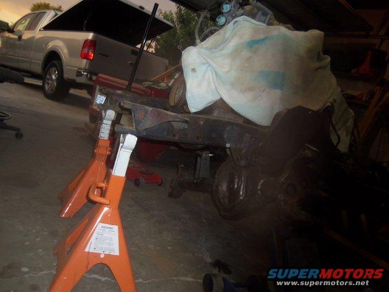 Vehicle frame and suspension components on jack stands, with a pickup truck in the background