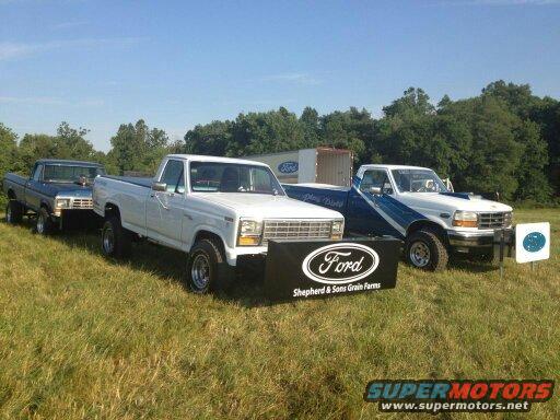 Three Ford trucks parked in a grassy field. A white 1980 Ford F-250 is in the foreground with a Ford banner. A blue Ford truck is behind it, and a white and blue Ford truck is to the right