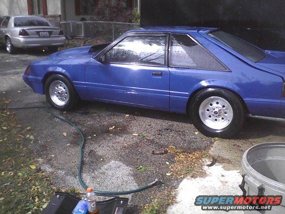 Blue coupe with aftermarket wheels and a hood scoop, parked on a wet driveway