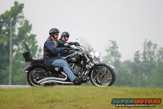 Two guys riding a black motorcycle on a road with grass and trees