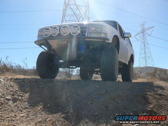 White SUV with off-road modifications, including large tires and Hella lights, driving on a dirt hill with power lines in the background
