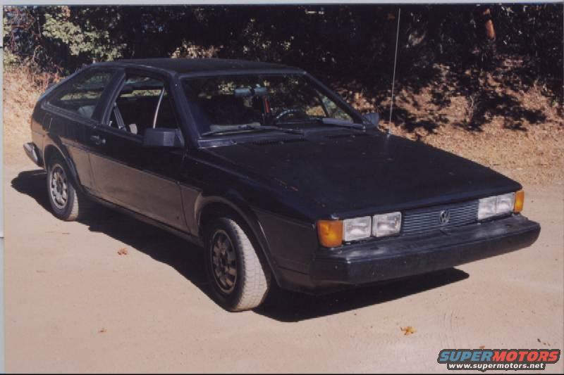 Dark hatchback car parked on a dirt road. It has square headlights and a grille with a VW emblem