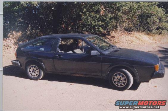 Dark blue Volkswagen Scirocco, side view, parked on dirt road with trees in background