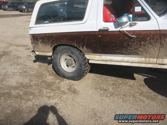 Side view of a muddy white and brown SUV, possibly a Ford Bronco, with a white camper shell. Other vehicles visible in background