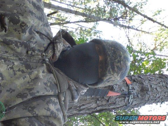 Camouflage-clad person wearing a black face mask and a fur-lined hat, sitting in a tree