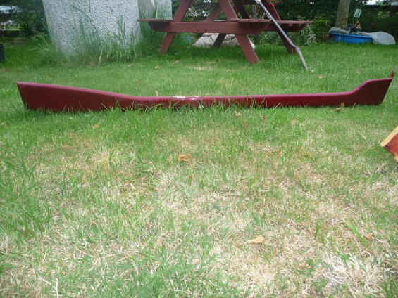 Red car side skirt lying on grass. The part is curved and appears to be made of plastic