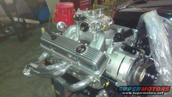 Close-up of a V8 engine with chrome valve covers, intake manifold, carburetor, and exhaust headers