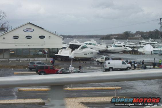 Boats washed ashore in a marina, with cars and vans nearby. A building is visible on the left