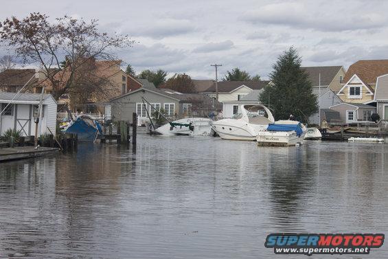Flooded canal with several boats docked and partially submerged, residential houses in the background under a cloudy sky