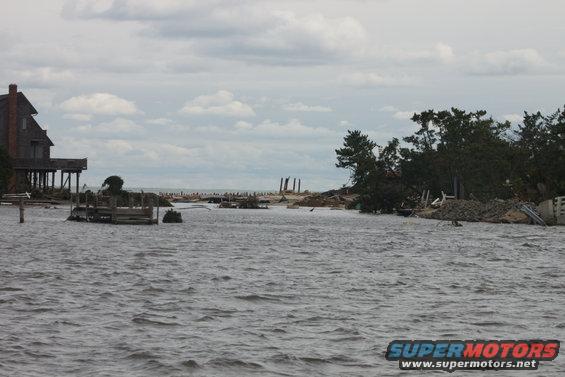 Flooded coastal scene with damaged house on stilts, debris, and trees under a cloudy sky