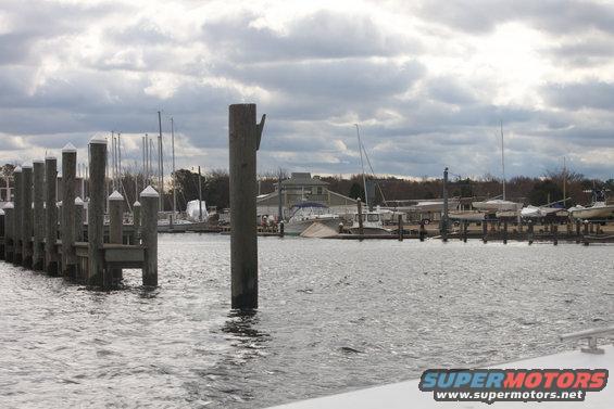 Marina with sailboats and motorboats docked under a cloudy sky. Wooden pilings line the left side