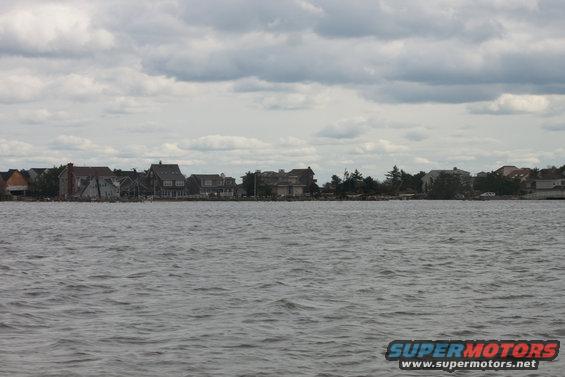 Waterfront houses under a cloudy sky, with a body of water in the foreground. Supermotors.net logo in the bottom right