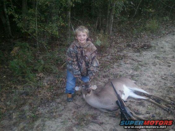 kid in camo jacket and jeans next to a dead deer with a rifle on the ground