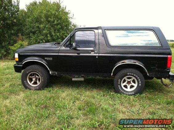 Side view of a black Ford Bronco parked on grass with trees in the background. The Bronco has chrome trim and aftermarket wheels