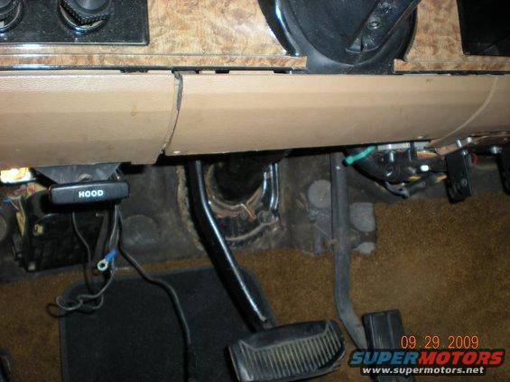 Interior view of a vehicle's dashboard with a hood release lever, pedals, and wiring visible