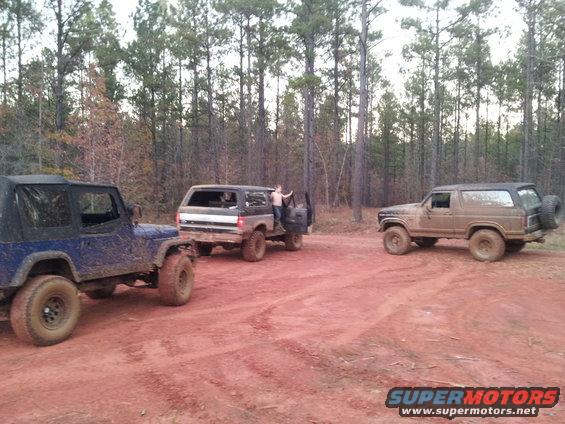 Three off-road vehicles parked on a dirt path in a forest. One is a blue Jeep, another is a dark SUV with a person standing in the open door, and the third is a tan SUV with a spare tire on the back