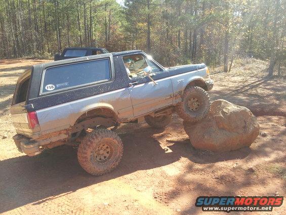 Muddy 1987 Ford Bronco climbing over a large rock on a dirt trail with another vehicle behind it