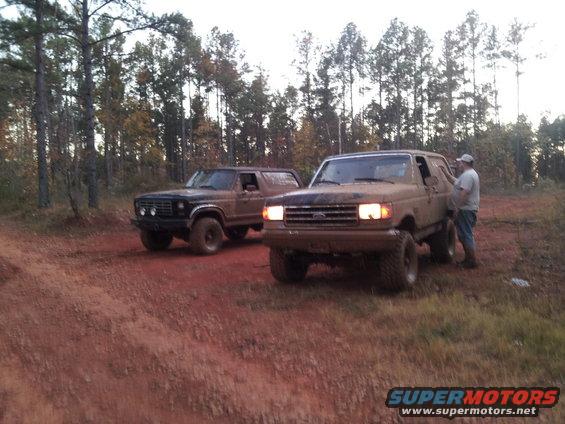 Two muddy Ford Broncos parked on a dirt road with trees in the background. The Bronco in the foreground is tan and the one behind it is dark