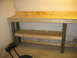 2004 Aston Martin Virage - A wooden workbench with a lower shelf and a black stool in front of it, set against a concrete wall.