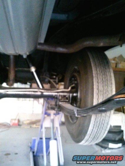 Underside view of a car's rear axle, leaf springs, and exhaust pipe. The vehicle is supported by jack stands