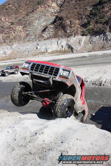 Red off-road truck with lifted suspension going through mud and sand. Another off-road vehicle is visible in the background