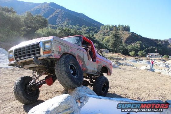 Red off-road truck with large tires climbing over rocks. Driver visible