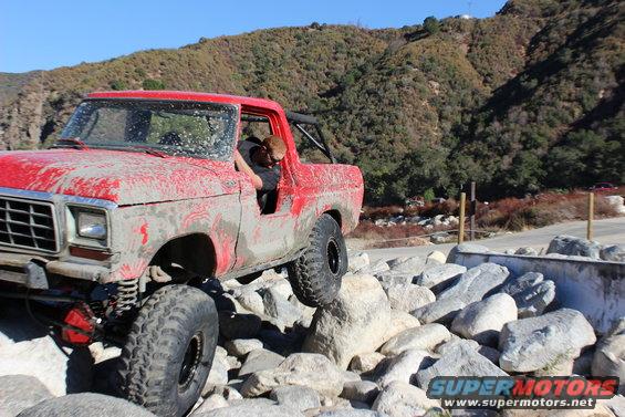 Red 1978 Ford Bronco with mud splatter, driving over rocks