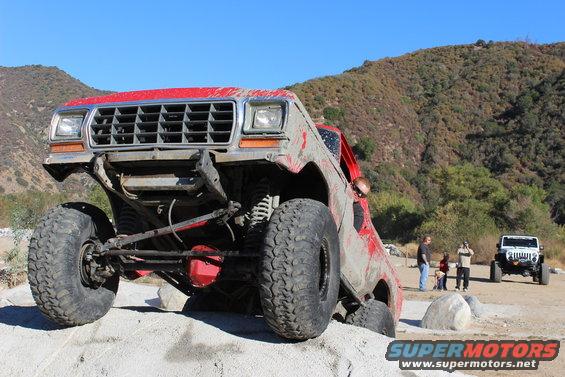 red off-road SUV with large tires, front suspension visible, on dirt terrain. Another white SUV is parked in background