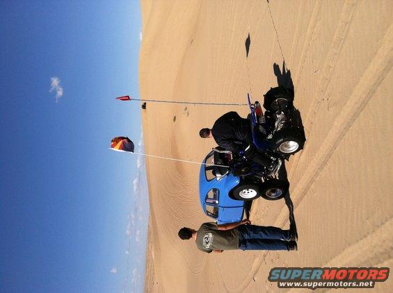 Blue dune buggy with two people on sand dunes, one person standing, one person sitting