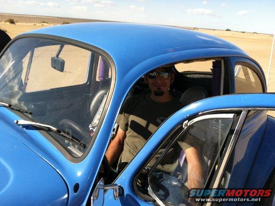 Blue 1970 Volkswagen Beetle with man driving, desert background. Supermotors logo visible